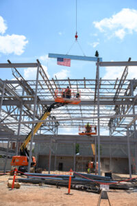 Workers on lifts assemble a steel frame structure at the Christ Church Episcopal School expansion site, with an American flag and a small tree fixed to a beam being lifted by Brasfield & Gorrie's crane.