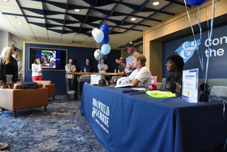 Varias personas se reúnen alrededor de una mesa decorada con globos y carteles de la empresa, mientras dos personas, sentadas, parecen participar en un acto de firma de contratos de trabajo de Brasfield and Gorrie en una sala moderna.
