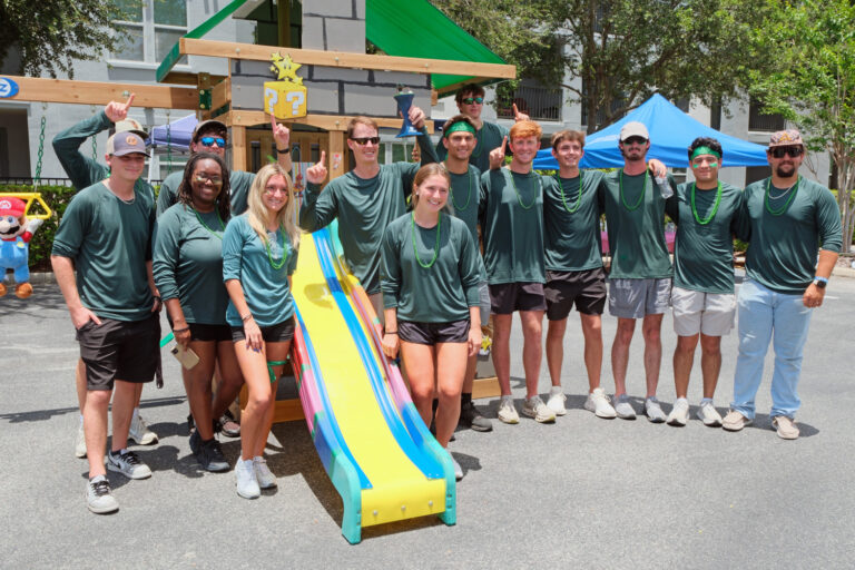 A large group of people in matching green shirts and bead necklaces pose in front of a playground structure while building custom playsets for children.