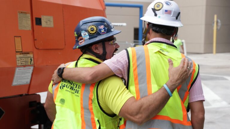 Two construction workers wearing safety vests and helmets stand side by side, one with his arm around the other's shoulder, near industrial equipment on a Brasfield and Gorrie jobsite.