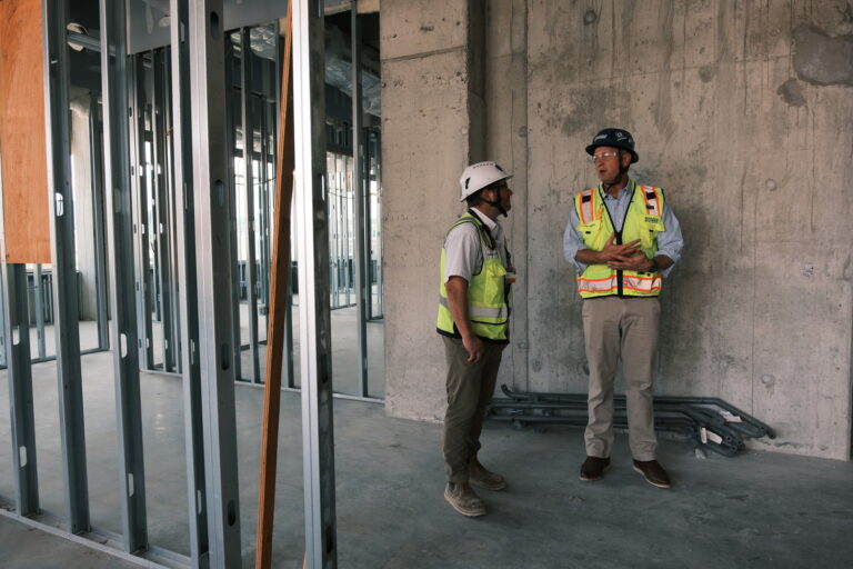 Two construction workers in safety vests and helmets stand and talk inside a building under construction, highlighting Brasfield and Gorrie's competitive advantage amid exposed concrete walls and metal framing.