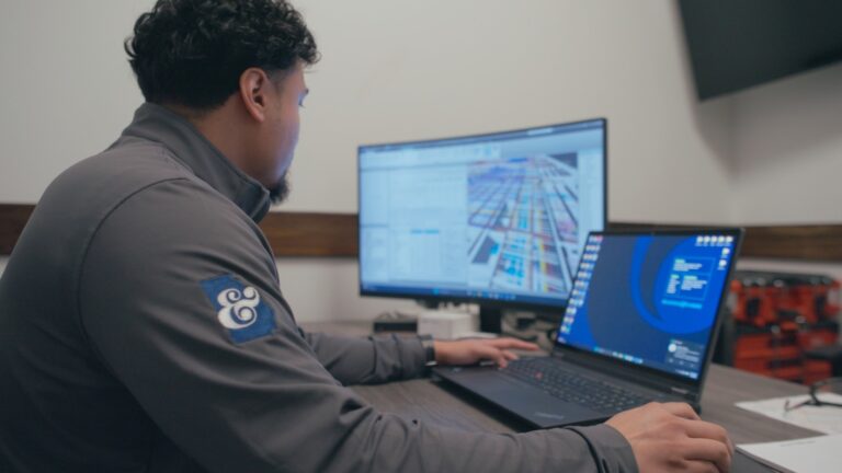 Un hombre trabaja en un escritorio con un ordenador portátil y un monitor en el que se ven diagramas técnicos y programas informáticos, en un entorno de oficina, mientras busca nuevas oportunidades profesionales en Brasfield and Gorrie.