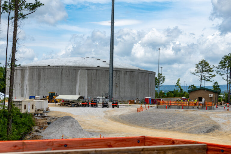 A large, round concrete water tank is surrounded by Chattanooga construction equipment, vehicles, and workers on a gravel lot with trees and a cloudy sky in the background.