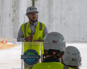 Una persona con casco y chaleco de seguridad habla desde un atril en el que se lee «La ciudad de Chattanooga, Tennessee», dirigiéndose a otros trabajadores de la construcción de Chattanooga reunidos en una obra en curso.
