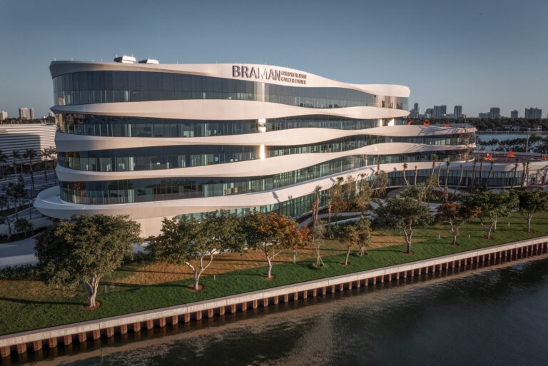 Modern, multi-story building with wavy glass facade, labeled “BRAMAN,” situated by the waterfront with a row of trees and city skyline in the background, home to Mount Sinai’s Braman Comprehensive Cancer Center.