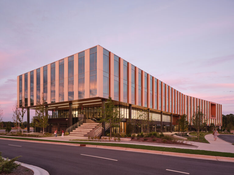 Modern building with large windows and vertical panels, set against a pink sky at the Raleigh Spark Campus, with landscaping, stairs, and a road in the foreground—a stunning project by Brasfield & Gorrie.