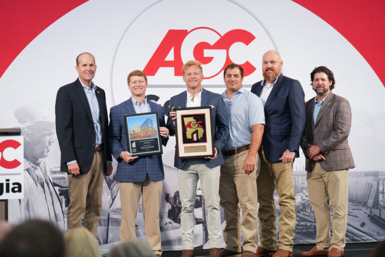 Six men in business attire stand on stage holding AGC Build Georgia awards plaques, posing for a group photo in front of an AGC-branded backdrop.