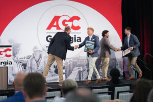 Four men in business attire shake hands and receive framed awards on stage at an AGC Georgia event, with a backdrop featuring the organization’s logo and celebrating the prestigious AGC Build Georgia awards.