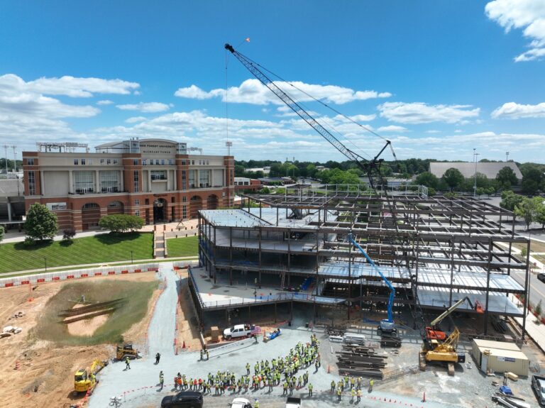 A construction site with a steel framework being built, a crane in use, workers gathered below, and the impressive Wake Forest University office building rising in the background under a blue sky with scattered clouds.