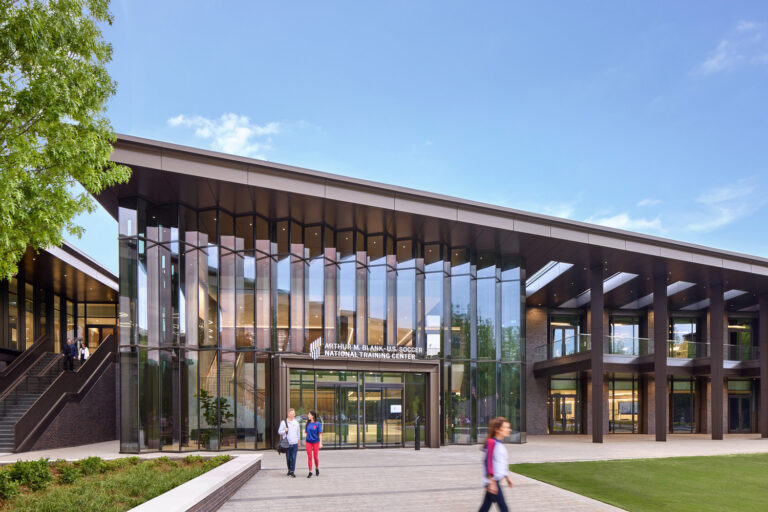 Modern glass building with large windows, labeled “National Training Center,” with people walking in front and a green lawn on the right side. This impressive U.S. Soccer Training Center provides a state-of-the-art environment for athletes honing their skills.