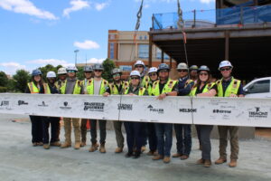 A group of construction workers in safety gear pose outdoors, holding a steel beam signed with various names and logos, at the Wake Forest University office building construction site.