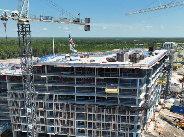A multi-story building under construction, with cranes, scaffolding, and an American flag on the roof. The site—part of the Fleet Landing Senior Living Community and built by Brasfield & Gorrie—is surrounded by trees and other buildings in the background.
