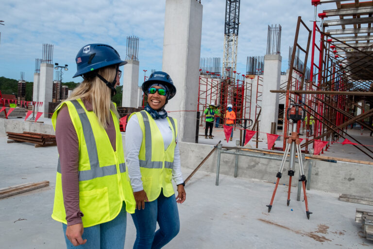 Dos mujeres, que llevan chalecos de seguridad y cascos, caminan y charlan en una obra en la que se ven andamios, columnas y trabajadores al fondo, lo que ilustra el espíritu de colaboración que se destaca en muchos de los «Brasfield and Gorrie Facts».