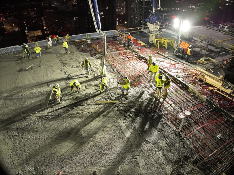 Los trabajadores de la construcción vierten y extienden hormigón en la azotea de un gran edificio por la noche, bajo la luz intensa de los focos de trabajo, con las barras de refuerzo y la maquinaria a la vista: una escena que recuerda a los grandes proyectos llevados a cabo por líderes del sector como Brasfield y Gorrie. Para quienes estén interesados en datos sobre Brasfield y Gorrie, sus equipos son conocidos por gestionar con maestría vertidos nocturnos complejos como este, con el fin de cumplir con los plazos y garantizar una calidad de primer nivel.