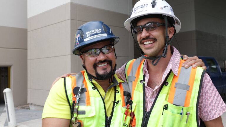 Dos trabajadores de la construcción, que llevan chalecos de seguridad, cascos y gafas de sol, sonríen abrazados delante de un edificio, representando con orgullo a Brasfield and Gorrie. Descubre más datos sobre Brasfield and Gorrie mientras observas su compromiso con el trabajo en equipo y la seguridad en cada proyecto.