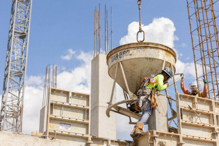 Un obrero de la construcción, equipado con material de seguridad, guía una cuchara de hormigón suspendida de una grúa en una obra en marcha, bajo un cielo azul salpicado de nubes, lo que pone de manifiesto la dedicación y la precisión que suelen caracterizar a los proyectos de Brasfield y Gorrie.