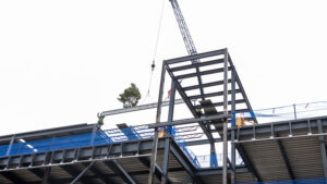A crane lifts a steel beam onto the Brasfield & Gorrie framework of the Wake Tech Simulation Hospital under construction, with two workers standing on the structure.