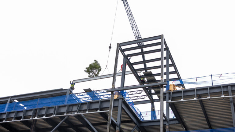 A crane lifts a steel beam onto the Brasfield & Gorrie framework of the Wake Tech Simulation Hospital under construction, with two workers standing on the structure.