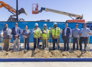 Nine people stand in a line at a Brasfield & Gorrie construction site for The Cade Hotel, holding ceremonial shovels in a pile of dirt with equipment in the background.