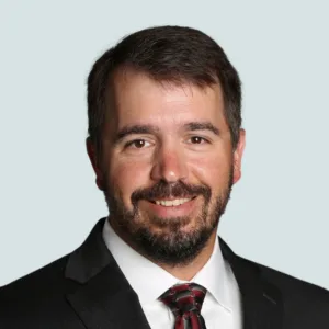 A man with a beard wearing a dark suit, white shirt, and patterned tie.