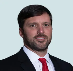 Headshot of a man with dark hair and a beard, wearing a black suit, white shirt, and red tie.