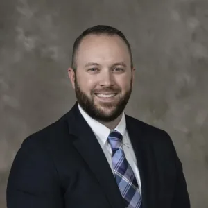 A man with a beard wearing a dark suit, white shirt, and plaid tie.