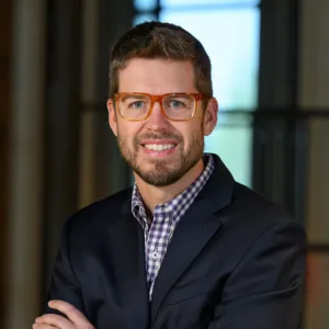 Michael Keller, a bearded man with orange-framed glasses, wears a navy blazer and a checkered shirt while posing with his arms crossed.