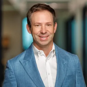 A man wearing a light blue blazer over a white button-down shirt, smiling at the camera.