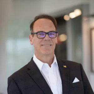 A man wearing glasses, a white collared shirt, and a dark blazer.