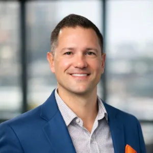Headshot of Mike Foushee smiling in a blue blazer and patterned dress shirt with an orange pocket square.