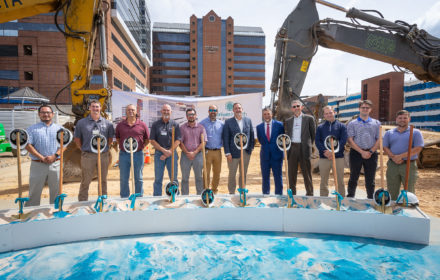 A dozen people stand behind shovels at a construction groundbreaking event