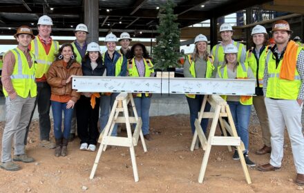 Thirteen people, mostly wearing hard hats and safety vests, gather around a signed steel beam topped by a small Christmas tree. The beam sits on a pair of plywood sawhorses. The group is gathered under a corrugated metal roof, and they are standing on red dirt.