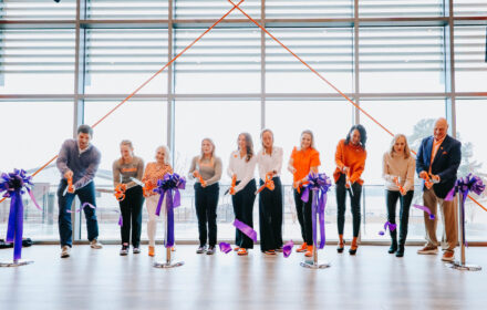 A row of 10 people, mostly women, stand in front of windows to cut a grand opening-type ribbon. The ribbon is purple and the people hold orange scissors--the colors of Clemson University.
