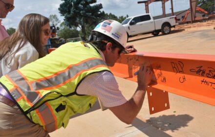 A man in personal protective equipment signs an orange steel beam on a construction site. Another man and a woman look on.