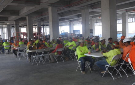 Construction workers in safety gear celebrating over lunch at tables inside a partially completed building