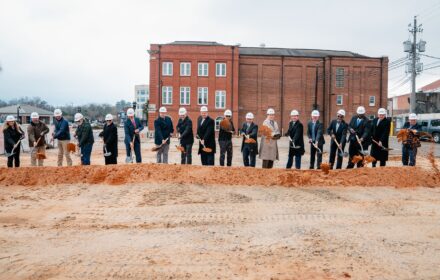 A line of 18 people in hardhats and holding shovels toss red dirt during a ceremonial groundbreaking