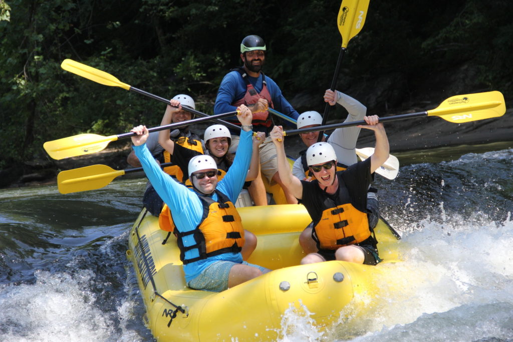 Team members white water rafting with their hands raised in the air