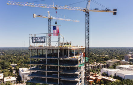 An aerial image of a steel beam being put in place at the top of a 26-story office building, with the city of Atlanta in the background