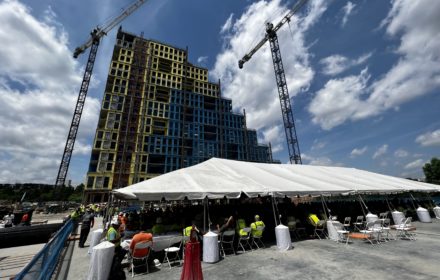 People gather under a white event tent in front of a residential tower under construction