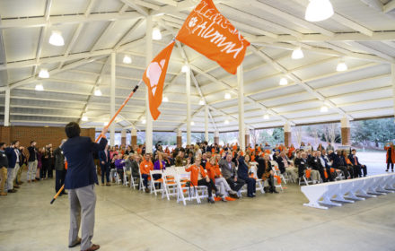 A man in a sportscoat and slacks waves an orange Clemson Alumni flag. An orange pawprint flag is on the pole beneath the alumni flag. A crowd of people in folding chairs look on.