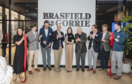 Nine people in business attire applaud behind a recently cut red ribbon. A woman in the center holds the ceremonial scissors. The group is inside of an office space, and Brasfield & Gorrie's logo is visible on the wall behind them.