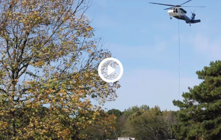 helicopter hovering above trees carrying HVAC equipment to install on a rooftop