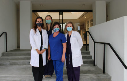 Four medical professionals wearing fabric face coverings gather in front of a medical office building