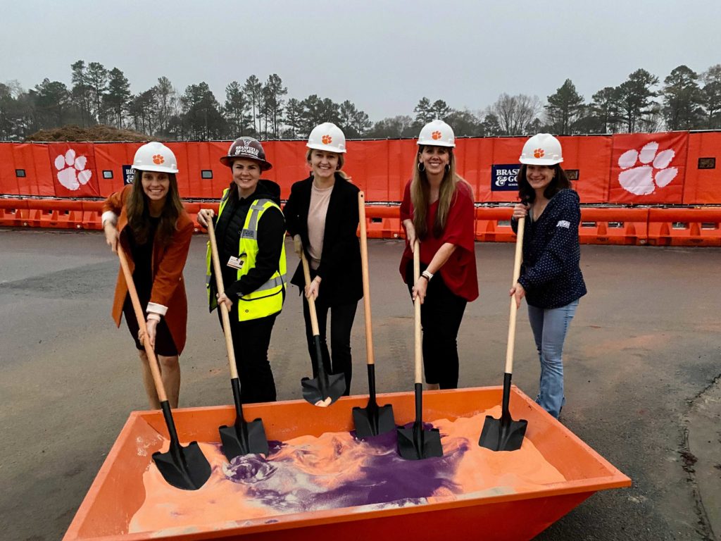 Five women in hard hats dig shovels into orange and purple sand during a groundbreaking event. They stand in front of jobsite fencing featuring the logos of Clemson University, which is a tiger paw print, and Brasfield & Gorrie, which is text. 