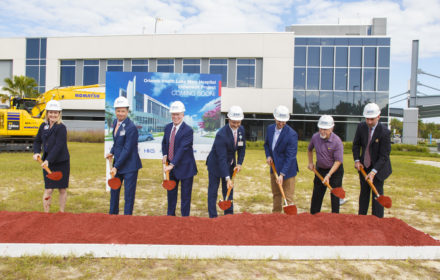 Seven people with shovels and hard hats break ground in front of a medical facility