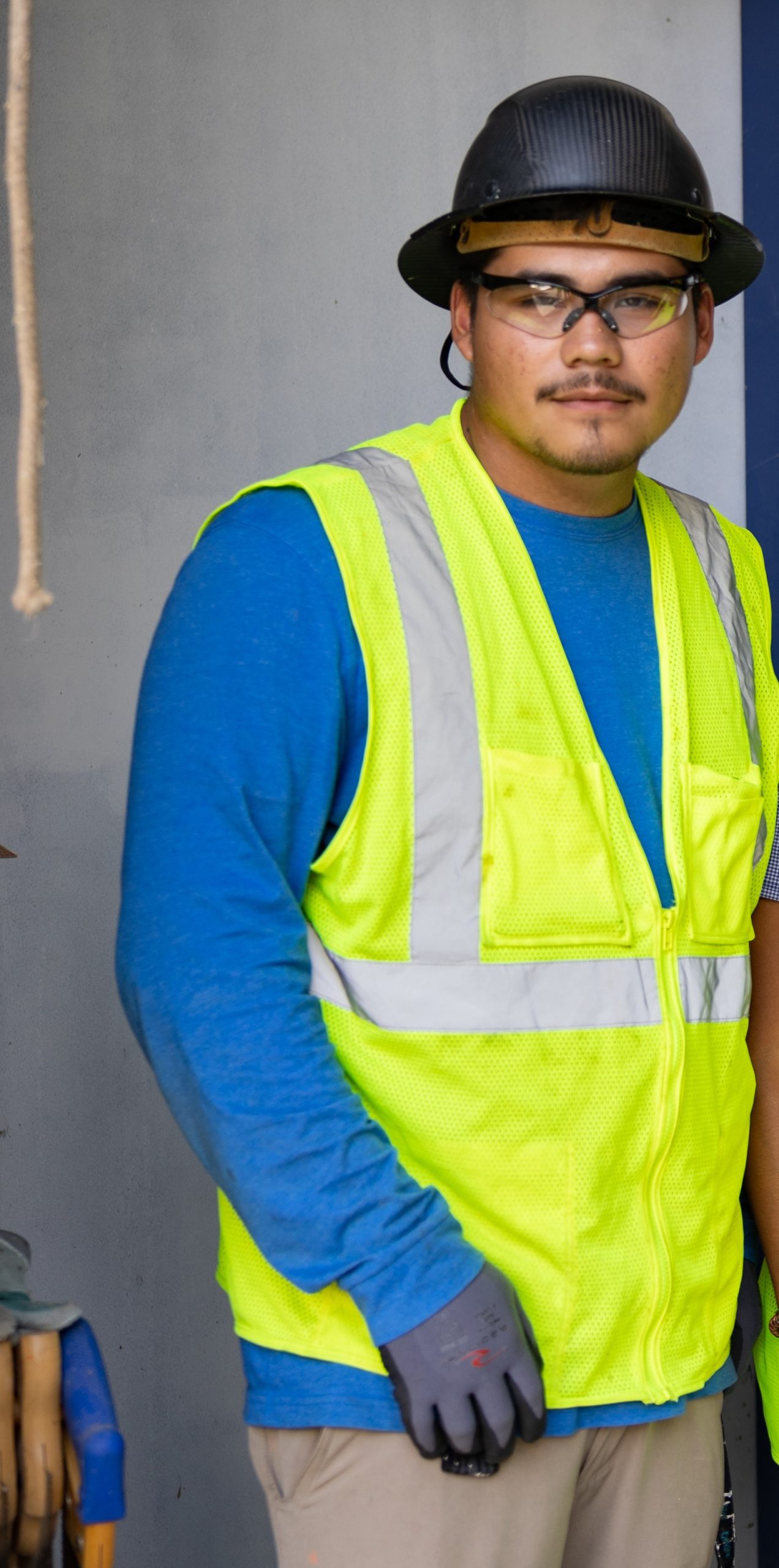 A male construction worker wearing a blue shirt and safety green vest with a brown hard hat