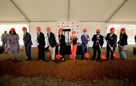 A group of people in business attire and hard hats scoop full of dirt at a groundbreaking event. The shovels and hard hats are red, to match Milo's Tea Company's branding. The group is gathered beneath a large event tent.