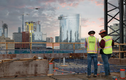 Two construction workers fly a drone at a construction jobsite with a city skyline in the background