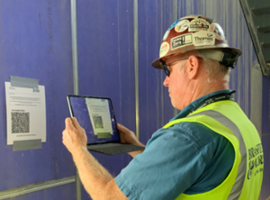 A male construction worker wearing personal protective equipment holds a tablet up to a QR code, which he will scan to access a digital dig board showing the construction site's utilities