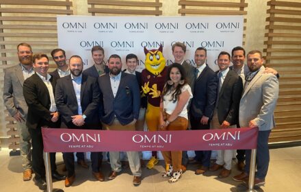 A group of 13 people gather with the Arizona State University mascot. The group is standing in front of an Omni Tempe at ASU backdrop and behind a ribbon that's marked with the same logo.